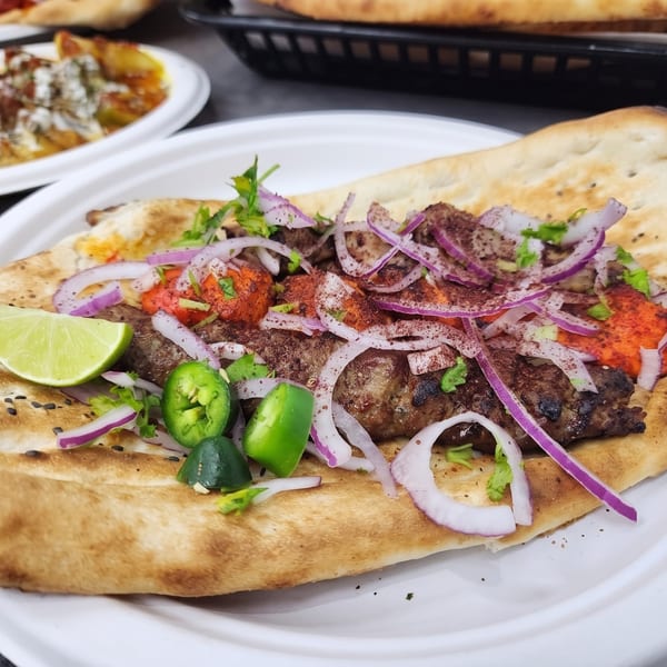 Grilled meat on flatbread with red onions, sumac, green peppers, cilantro, red sauce, and lime.